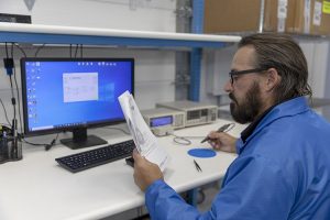 An employee at AGS Devices holding tweezers for electronic components and examining a document in a close-up view.